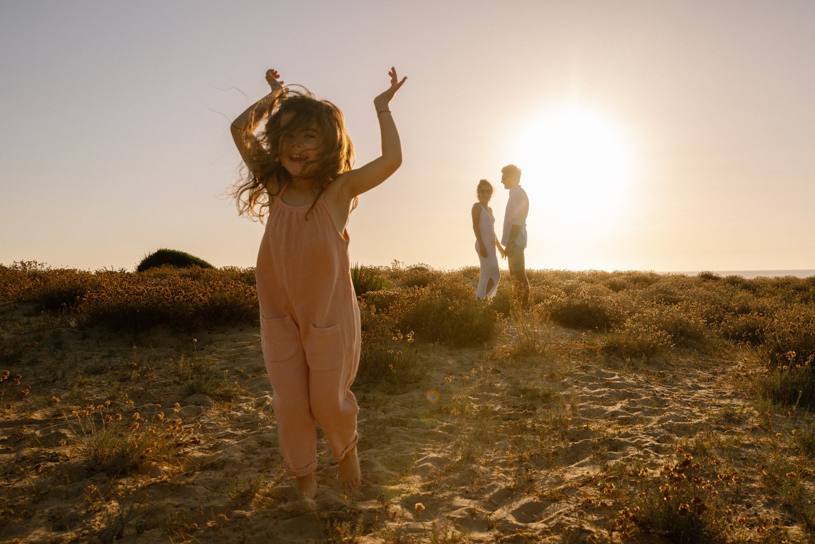 Une petite fille joyeuse saute au premier plan sur une plage de dunes au coucher du soleil, avec ses parents flous en arrière-plan sous une lumière dorée.